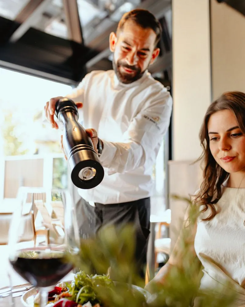 Chef grinding pepper over a plate while a guest watches